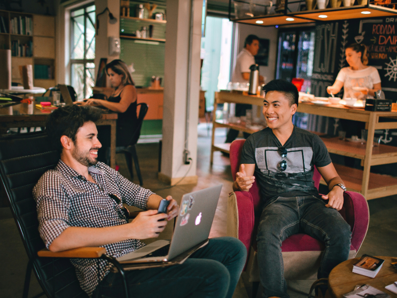 Two young professionals discussing work, one with a laptop, smiling in a casual office environment, illustrating interpersonal communication—a key MBA skill.