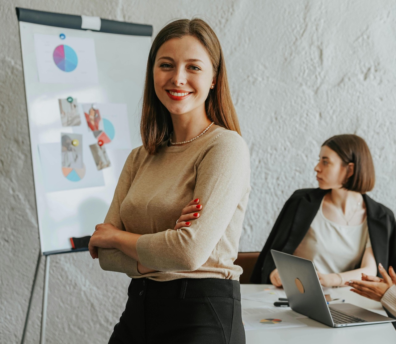 A confident professional stands in the foreground of a modern office, symbolizing career advancement. In the background, two colleagues engage in discussion at a table, with a flip chart displaying pie charts and notes, representing strategic marketing planning and career growth.